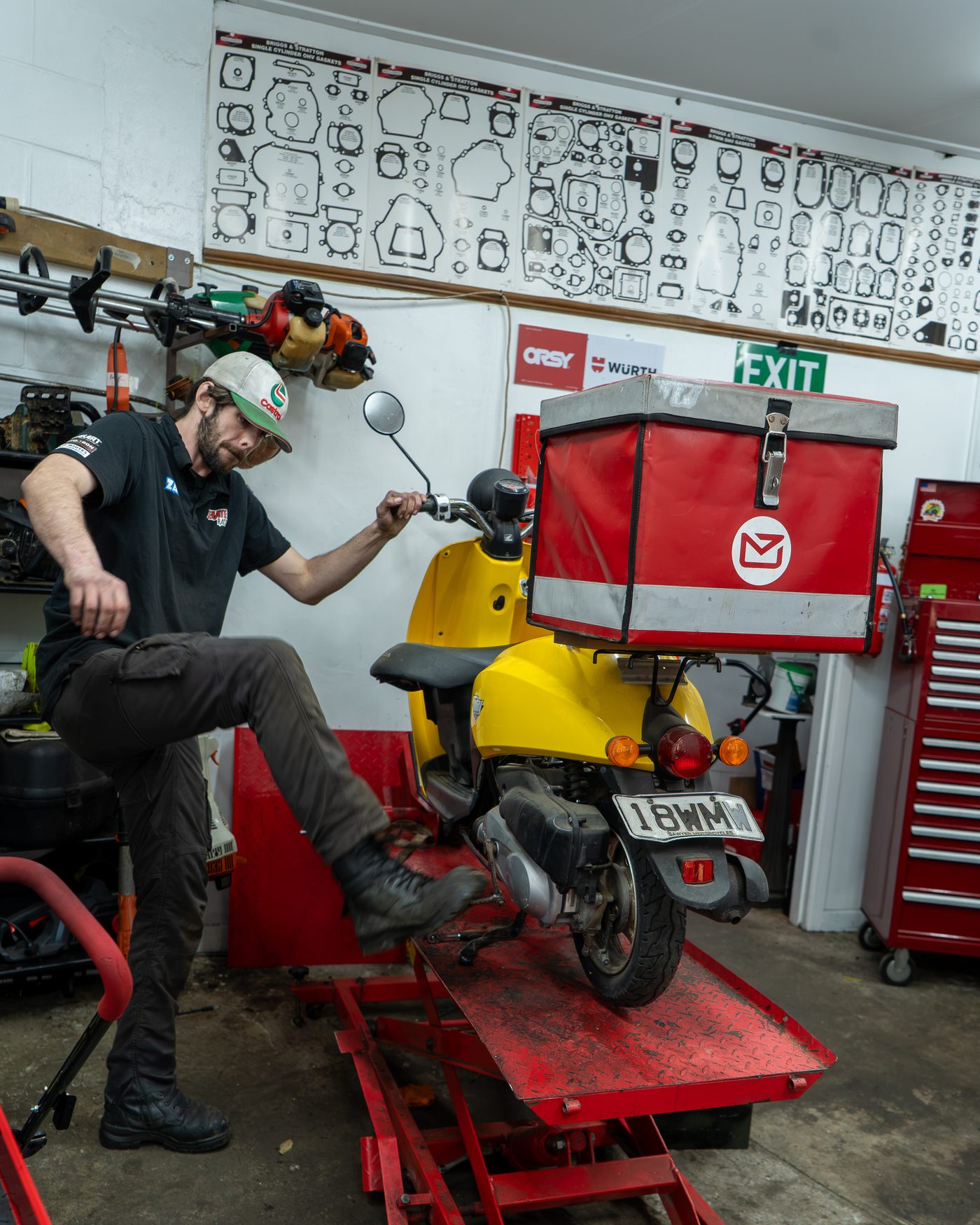 Mechanic kicking a scooter up onto a service lift in the workshop.